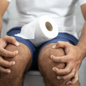 man on toilet grabbing his knees with a roll of toilet paper on his lap.